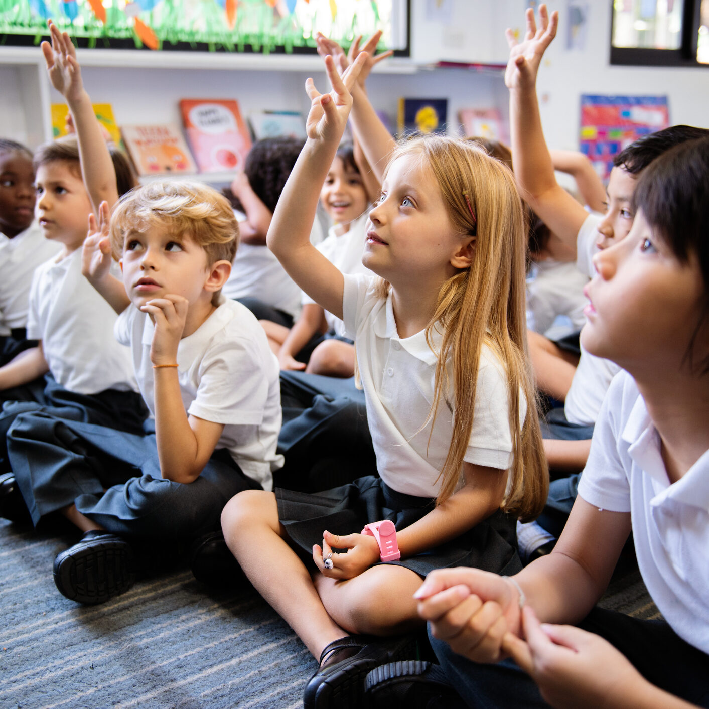 Diverse Group Of Young Students, Boys And Girls, Sit On The Floor In A Classroom Raising Hands.
